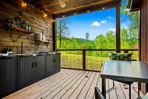 A wooden deck with a kitchen area, including cabinets and a table, overlooking a green landscape.