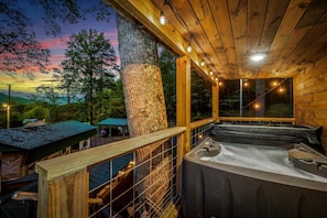 A wooden cabin with a hot tub, a covered carport, and a view of the surrounding forest.