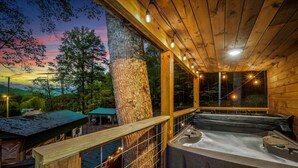 A wooden cabin with a hot tub, a covered carport, and a view of the surrounding forest.