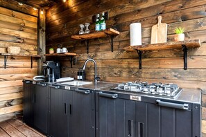 A compact kitchen with a sink, a coffee machine, a gas stove, and wooden shelves with various items. - Beautiful Cabin with Fire Pit in Hayesville, North Carolina (Hayesville)