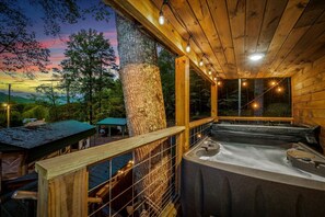 A wooden cabin with a hot tub, a covered carport, and a view of the surrounding forest.
