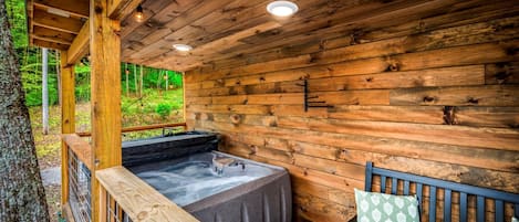 A wooden cabin with a hot tub, a bench, and a railing overlooking a forest.
