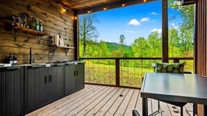 A wooden deck with a kitchen area, including cabinets and a table, overlooking a green landscape.