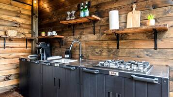 A compact kitchen with a sink, a coffee machine, a gas stove, and wooden shelves with various items.