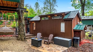 A wooden cabin with a patio, two Adirondack chairs, a fire pit, and a trash can.