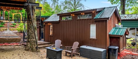 A wooden cabin with a patio, two Adirondack chairs, a fire pit, and a trash can.