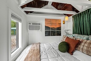 A bedroom with a bed, a window, an air conditioning unit, a wall-mounted lamp, and a view of mountains.