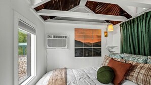 A bedroom with a bed, a window, an air conditioning unit, a wall-mounted lamp, and a view of mountains.