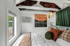A bedroom with a bed, a window, an air conditioning unit, a wall-mounted lamp, and a view of mountains.