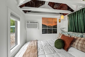 A bedroom with a bed, a window, an air conditioning unit, a wall-mounted lamp, and a view of mountains.