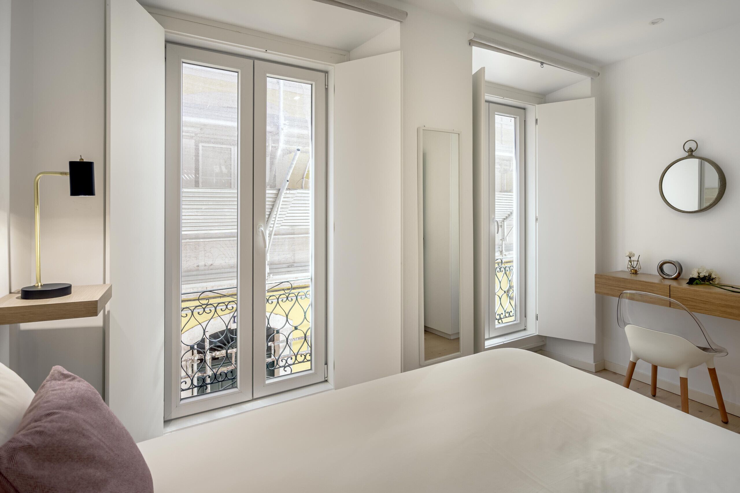 A bedroom with a bed, a desk, a chair, a mirror, and a window with a view of a balcony.