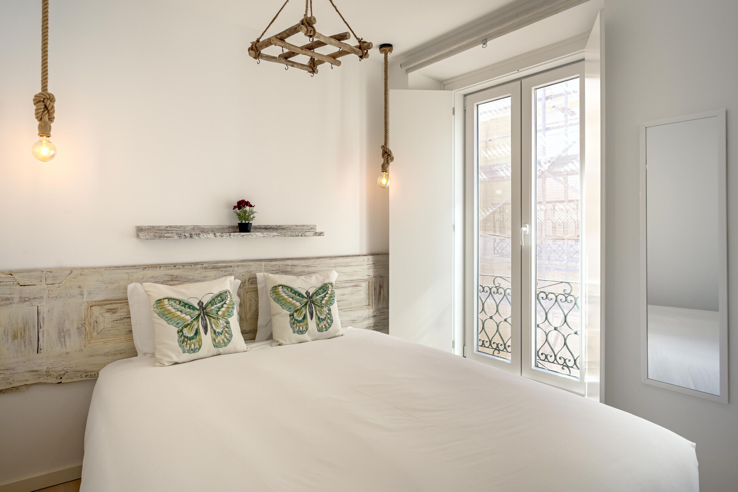 Ein Schlafzimmer mit einem großen Bett, zwei Kissen mit Schmetterlingsmuster, einem Holz-Kopfteil, einer kleinen Ablagefläche mit einer Pflanze und einem Fenster mit einem Metallgitter.
