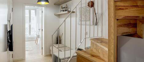 Un escalier moderne avec des marches en bois et des rampes en métal, menant à une chambre à coucher équipée d’un lit et d’une table de chevet.