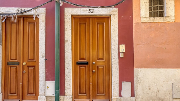 Zwei Holztüren mit der Nummer 52 darüber, eingebaut in ein Gebäude mit einer steinernen Basis und einem gepflasterten Vorplatz.