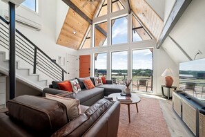 A modern living room with a large sectional sofa, a coffee table, and a TV. There is a staircase with a railing leading to an upper level.