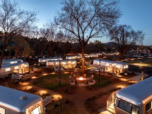 Un camping con vehículos recreativos, un área central con fogata y un árbol en el centro.