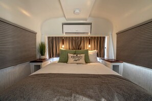 Une chambre à coucher avec un lit, une table de chevet, une plante en pot, un oreiller orné d’un motif et un appareil de climatisation.