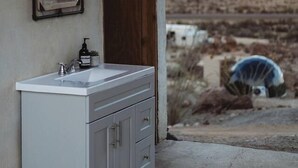 Ein Badezimmer mit einem weißen Waschbecken, einem Spiegel und Blick auf eine Wüstenlandschaft durch eine offene Tür.
