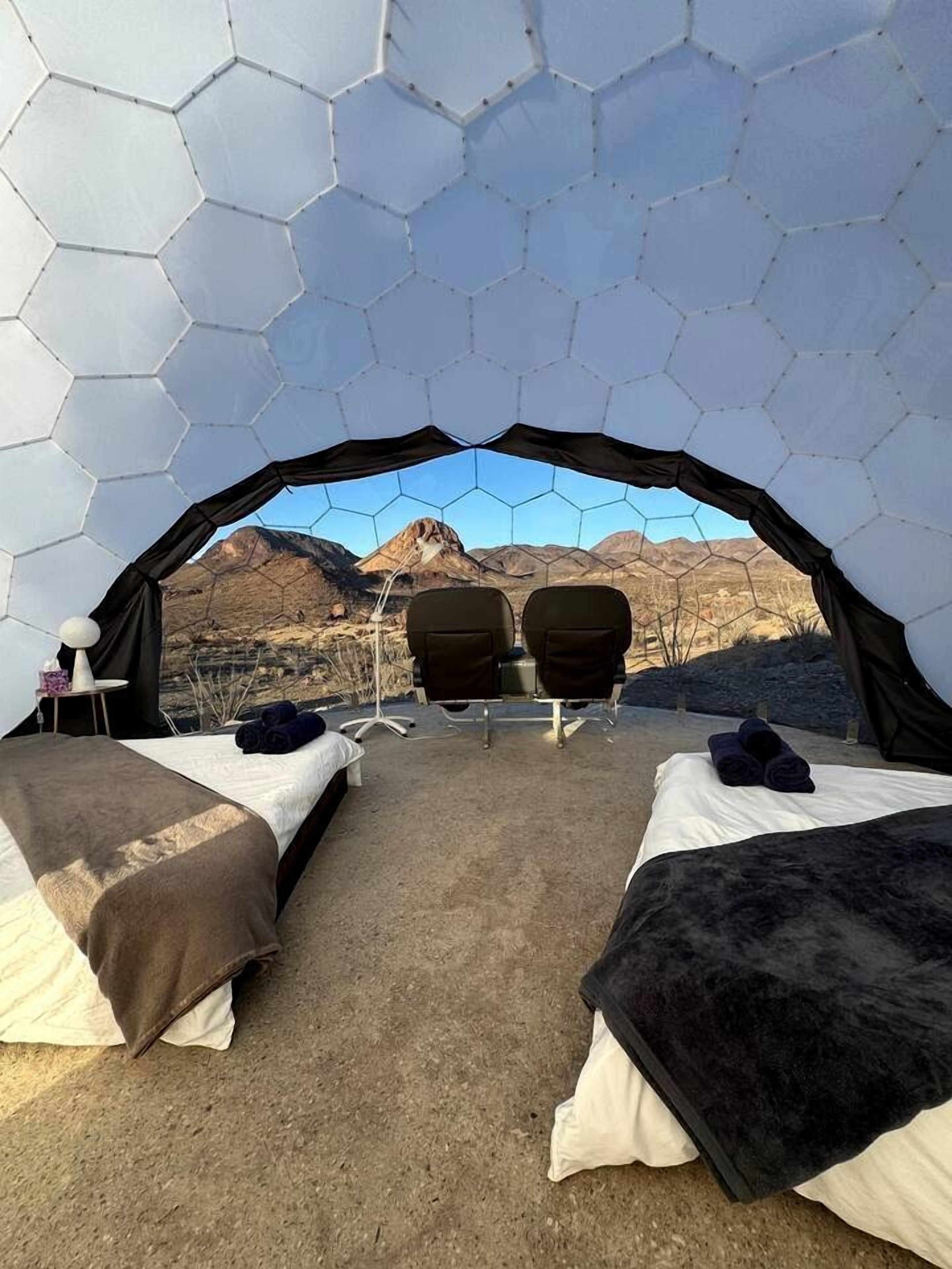 Une tente hexagonale comprenant deux lits, une table et des chaises, installée dans un décor de montagnes.