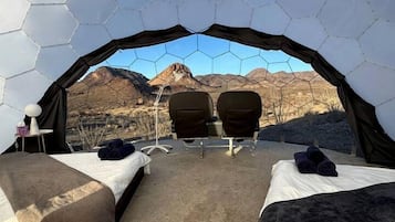 Une tente hexagonale comprenant deux lits, une table et des chaises, installée dans un décor de montagnes.