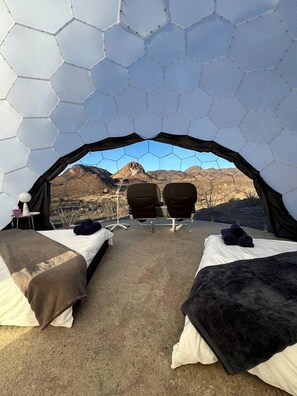 Une tente hexagonale comprenant deux lits, une table et des chaises, installée dans un décor de montagnes.