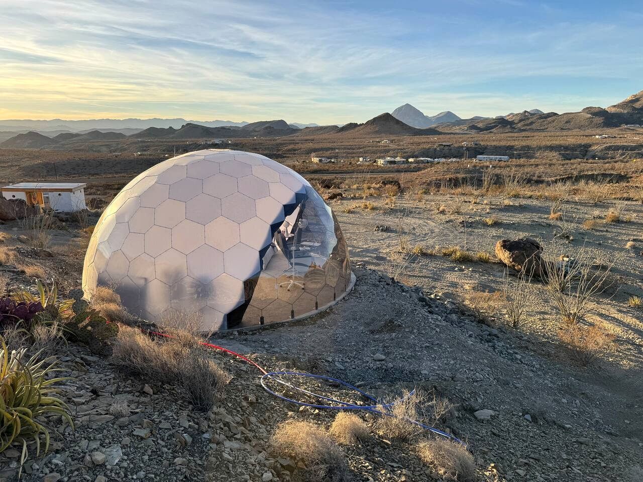 Une structure en forme de dôme géodésique, située dans un paysage désertique, avec des montagnes en arrière-plan.