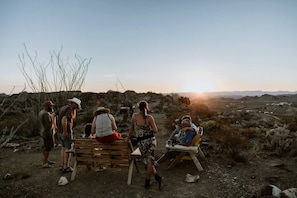 Un grupo de personas al aire libre, con un banco y sillas de madera, cielo despejado y un atardecer de fondo.