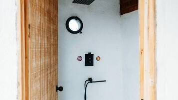 A small, enclosed bathroom with a wooden door, a white wall, a circular light fixture, a black electrical outlet, and a black hose.