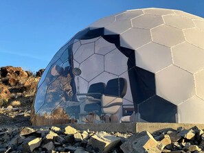 A geodesic dome structure with a reflective interior, a seating area, and a rocky outdoor environment.