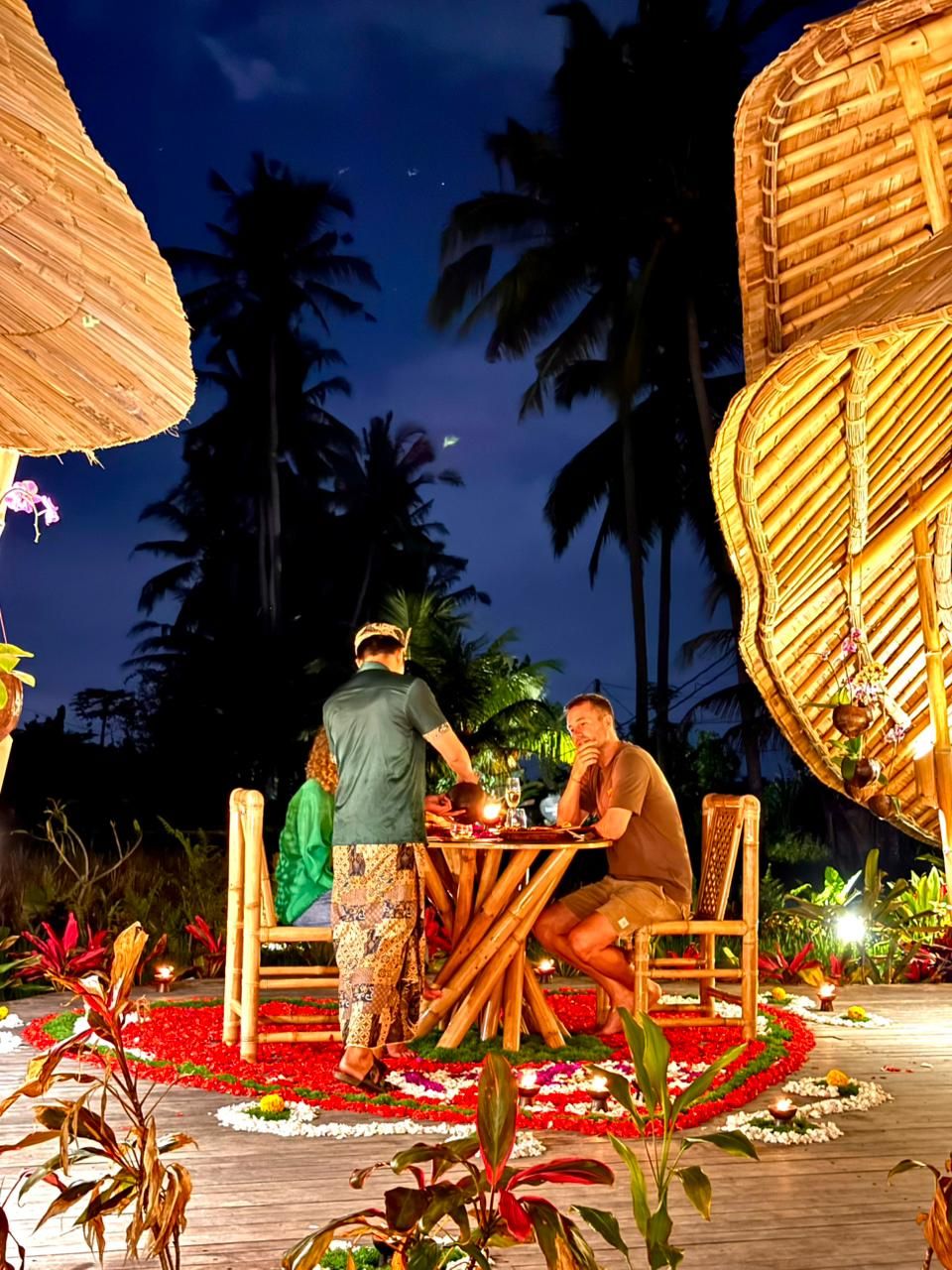A couple dining at an outdoor table with a thatched roof, surrounded by tropical plants and flowers.