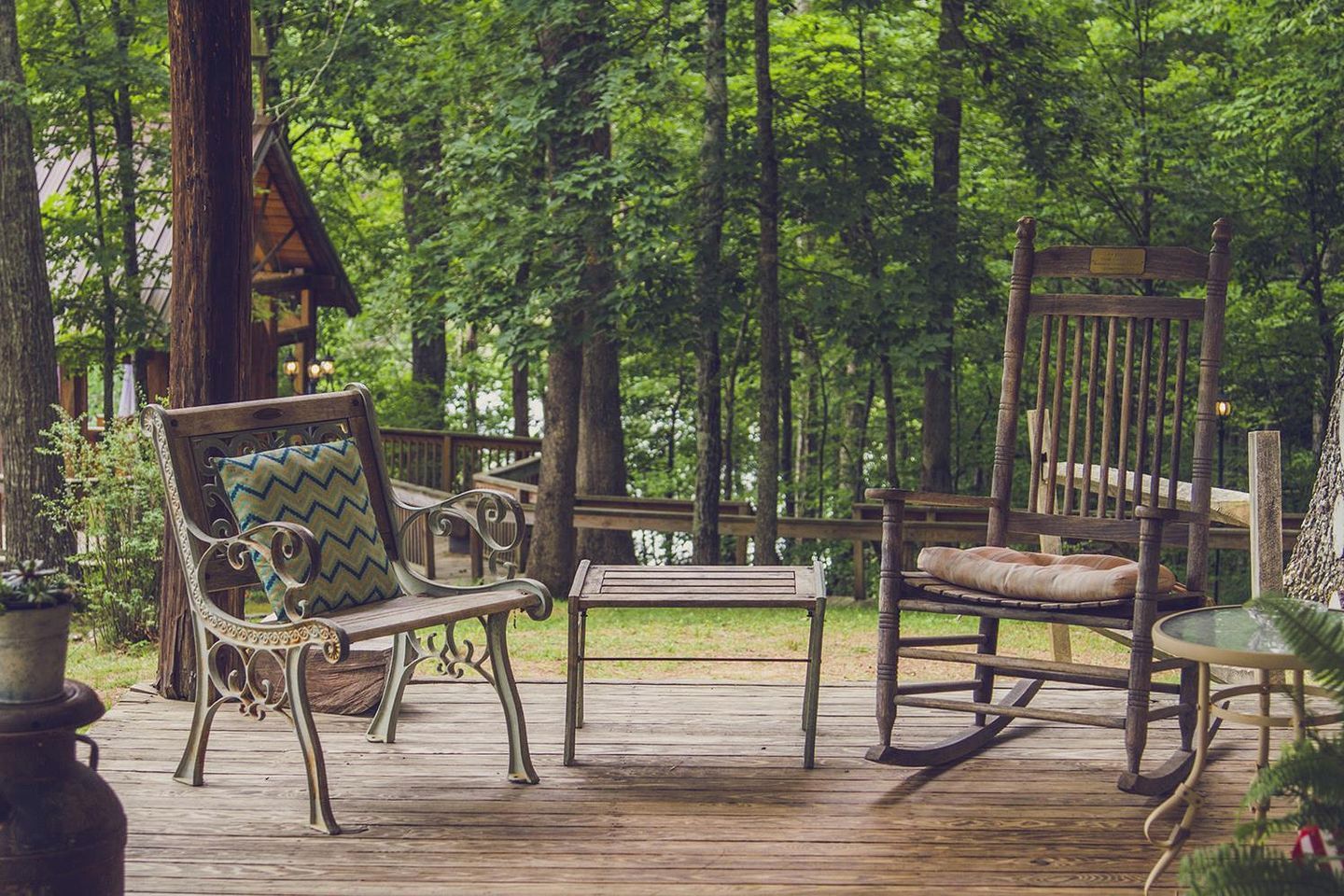 Une terrasse en bois avec un banc, une chaise berçante et une petite table, le tout entouré d’une zone boisée.