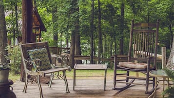 Une terrasse en bois avec un banc, une chaise berçante et une petite table, le tout entouré d’une zone boisée.