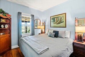 Une chambre à coucher comprenant un lit, une table de chevet, une lampe, une commode, une fenêtre avec des rideaux et un tableau encadré accroché au mur.