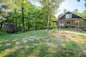 Une cabane rustique en rondins de bois, dotée d’un porche et entourée d’une zone boisée.