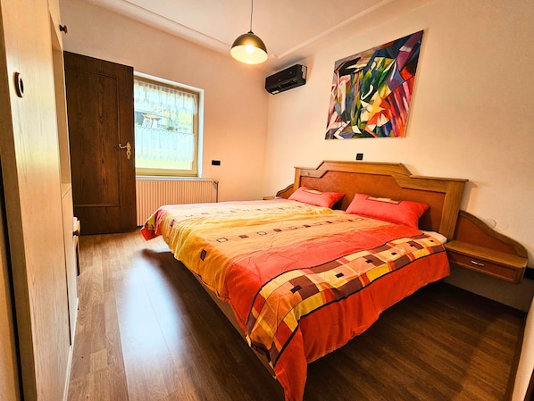 A bedroom with a wooden bed, a window with a view, a ceiling fan, and a painting on the wall.