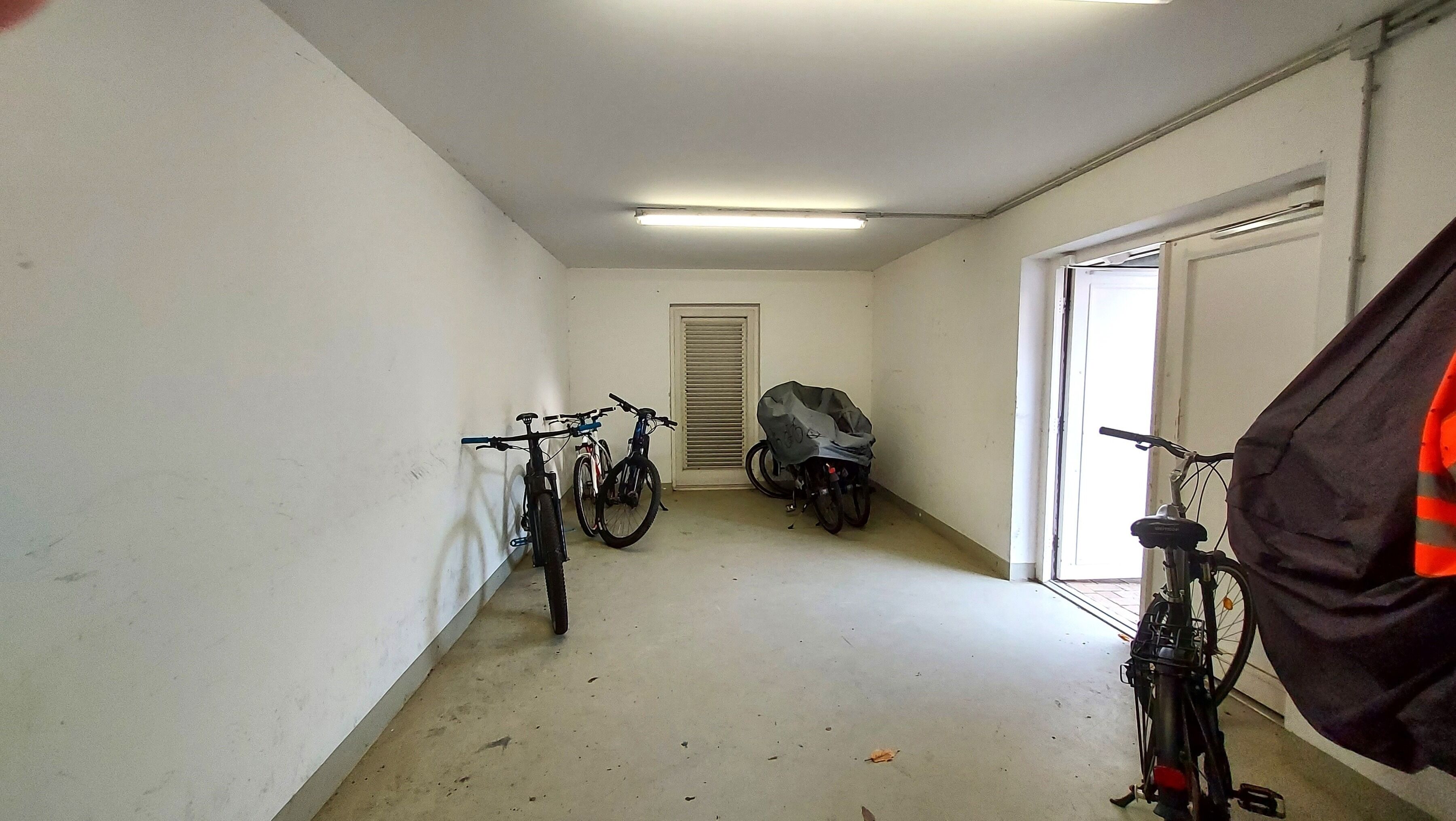 Ein schmaler Flur mit Fahrrädern an den Wänden, einer Tür am Ende und einem Fenster zur rechten Seite.