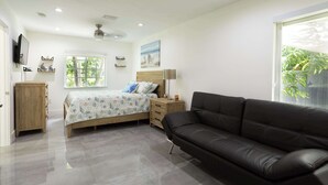 Un dormitorio moderno con cama, sofá de cuero negro, mesita de noche de madera, televisor y ventana con vistas a árboles.