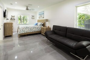 A modern bedroom with a bed, a black leather sofa, a wooden nightstand, a TV, and a window with a view of trees. - Tropical Paradise | Boat Parking | Pet Friendly (Miami)