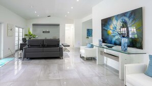 Una sala moderna con un sofá gris, sillones blancos y una mesa de centro de vidrio. Se aprecia un cuadro abstracto de gran tamaño en la pared y un florero sobre la mesa.