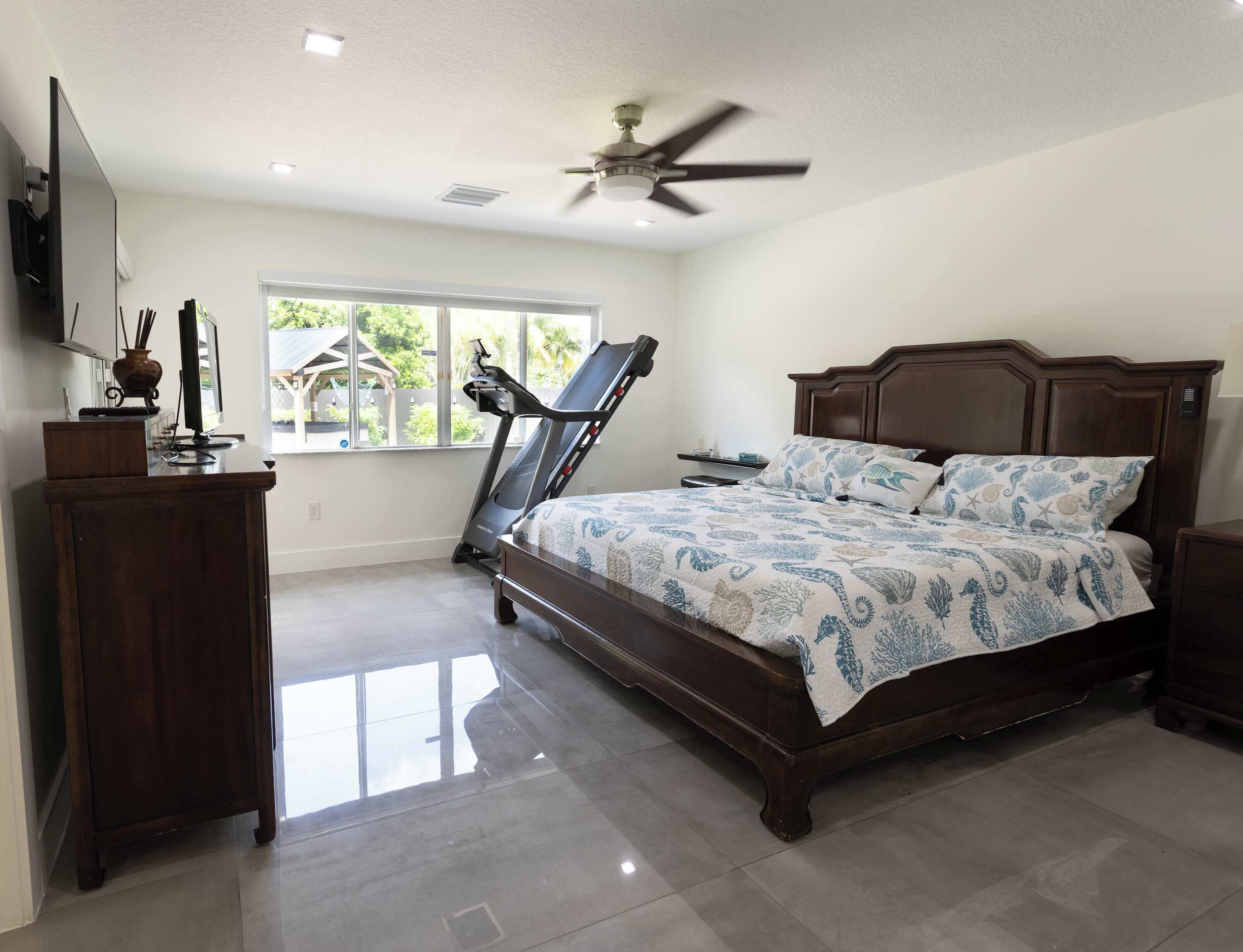 A bedroom with a bed, a treadmill, a desk, and a window with a view of greenery.
