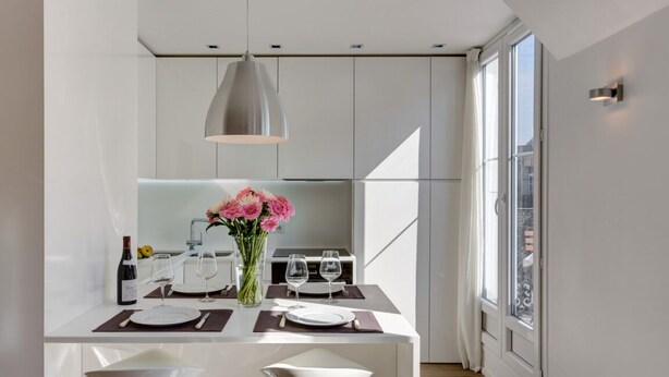 A modern kitchen with white cabinetry, a dining table set for two, and a large window providing natural light. - CHAMPS ELYSEES APARTMENT (Paris)