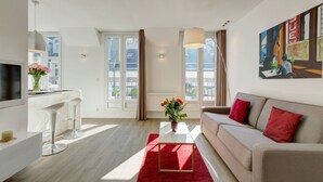 A modern living room with a sofa, a coffee table, and a TV. There are large windows with curtains, a painting on the wall, and a vase with flowers on the coffee table. - CHAMPS ELYSEES APARTMENT (Paris)