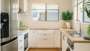A modern kitchen with stainless steel appliances, white cabinetry, a built-in washing machine, and a window with a view of the outside. - A Sunday Kind of Stay -  Home Near Manly + Shops (Capalaba)
