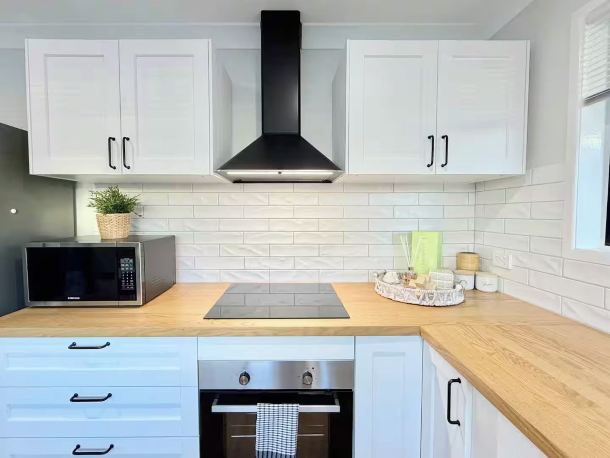 A modern kitchen with white cabinets, a black range hood, a microwave, a built-in oven, and a wooden countertop.