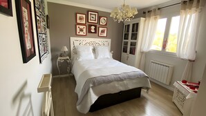 Une chambre à coucher avec un lit, une table de chevet, une commode, un radiateur et une fenêtre avec des rideaux.