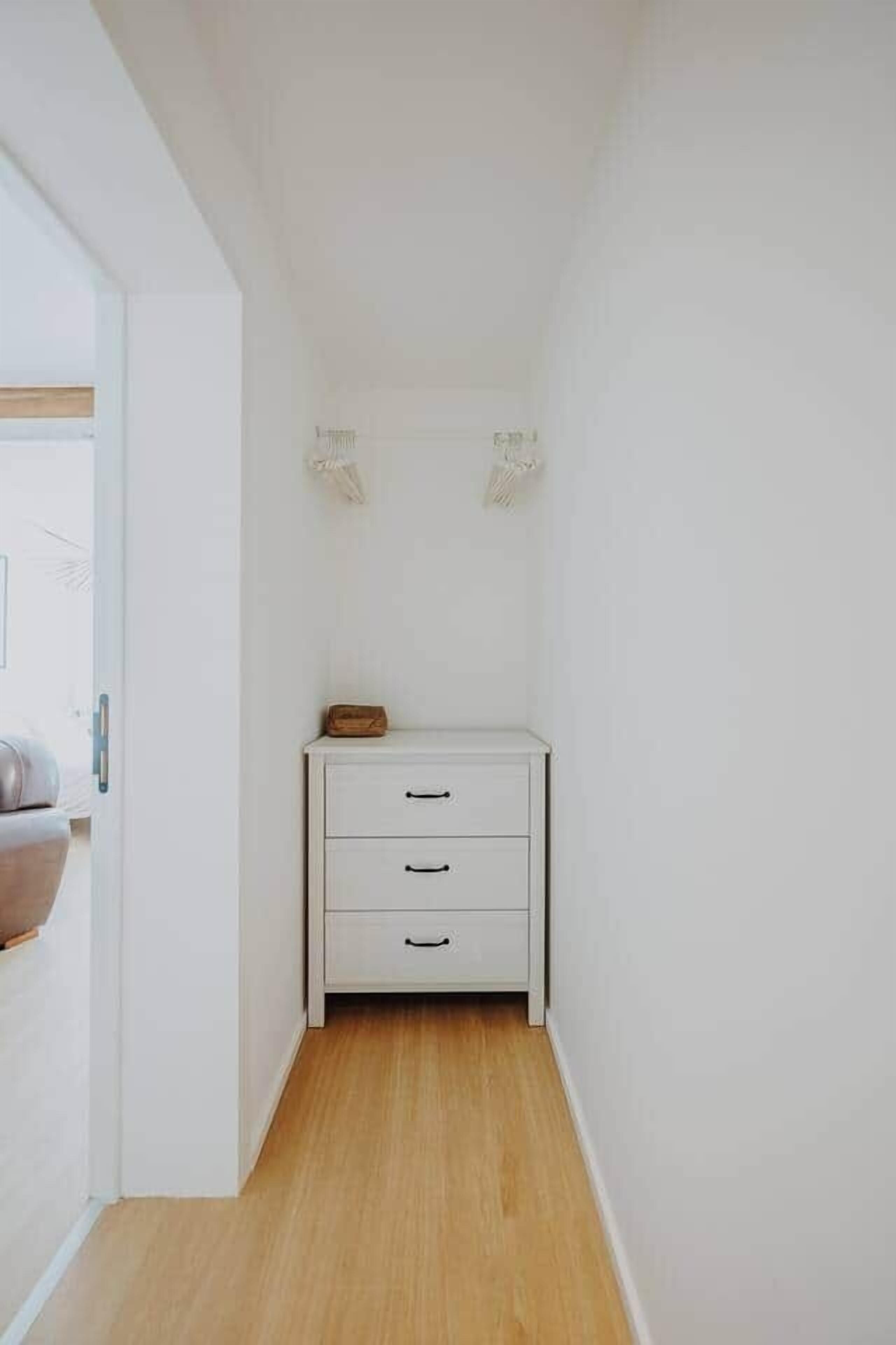A narrow hallway with a white wall and wooden floor, leading to a small white cabinet with drawers.