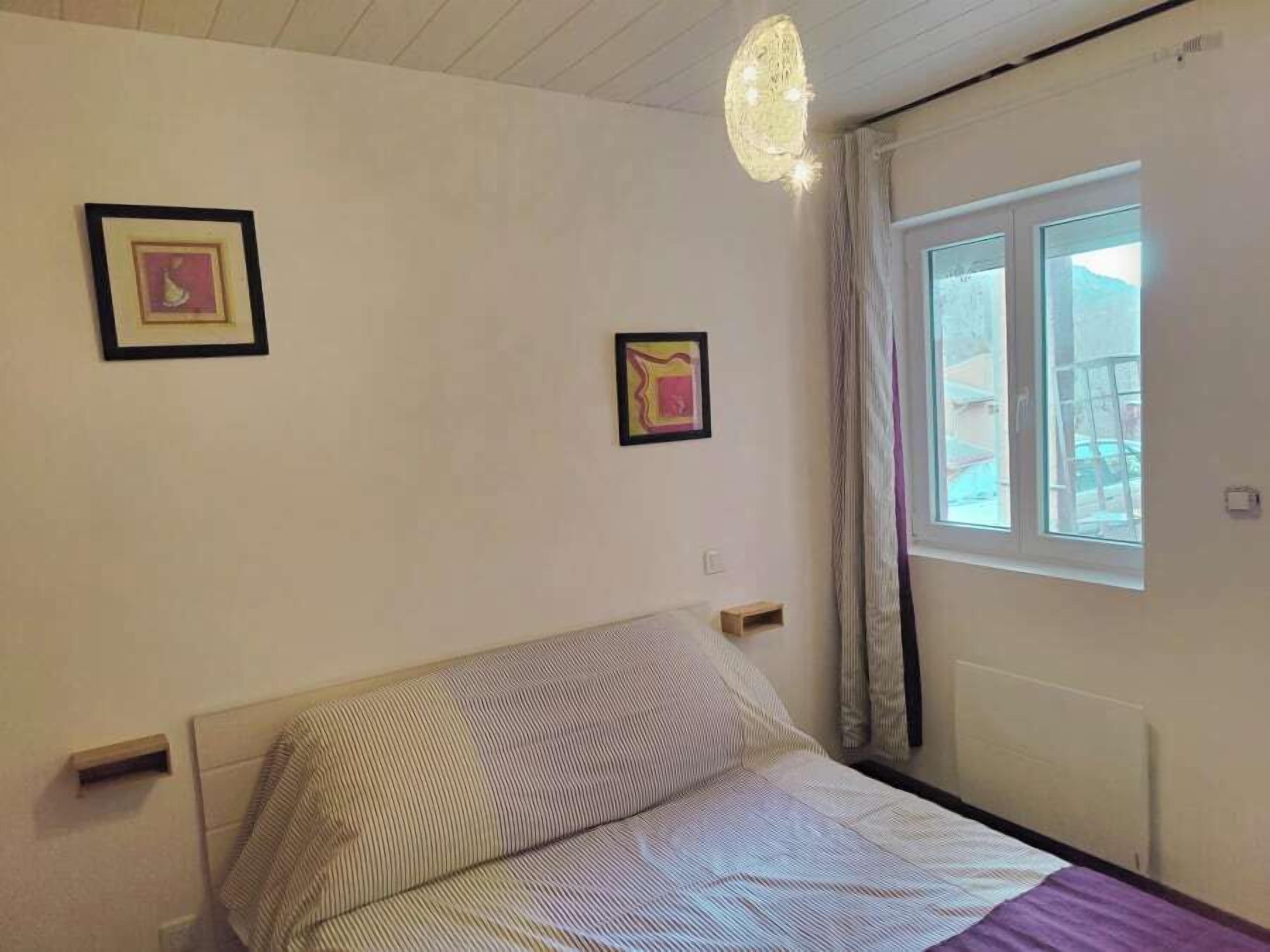 A bedroom with a bed, two framed pictures on the wall, a window with curtains, and a ceiling light fixture.