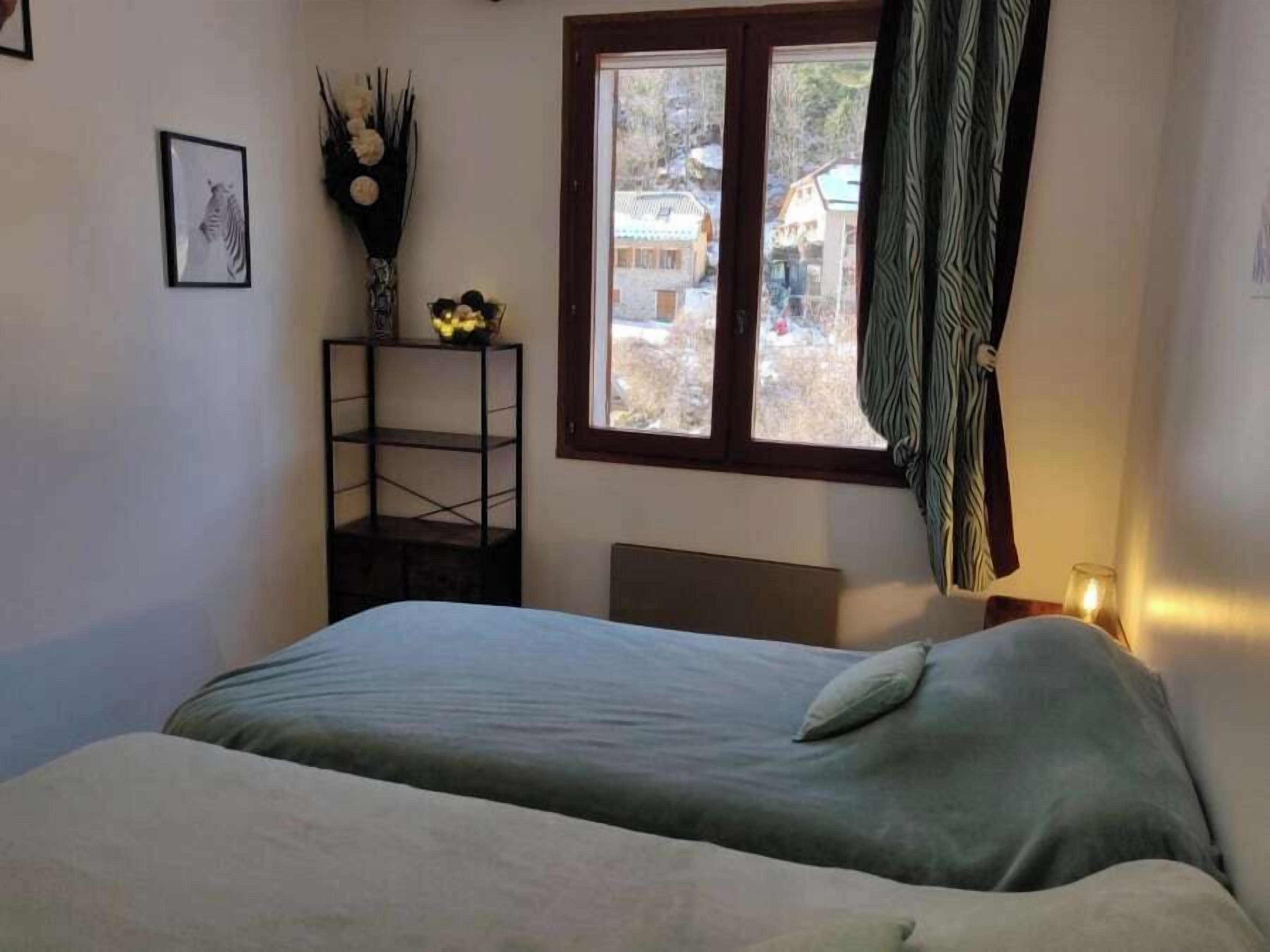A bedroom with a bed, a window with a view of buildings, a shelf with flowers, and a framed picture on the wall.