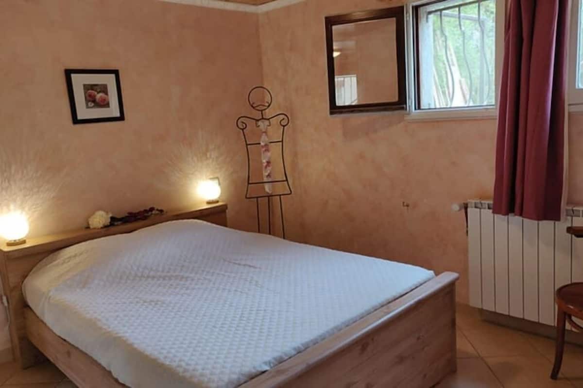 Un dormitorio con una cama de madera, una mesita de noche con lámpara, un cuadro en la pared y una ventana con cortinas.