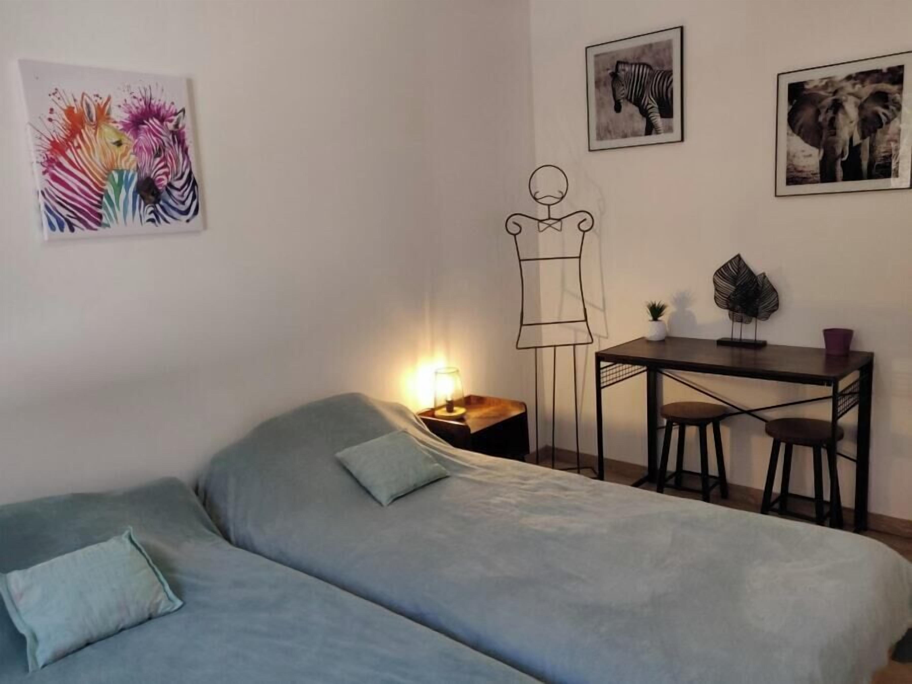 A room with a bed, a desk with two chairs, a small table with a plant, and two framed pictures on the wall.